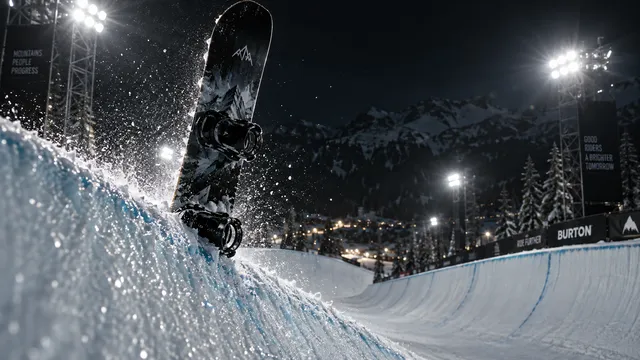 4K 16:9 widescreen action-still cinema shot at a mountain terrain park at night, intense halogen stadium floodlights backlighting a freestyle snowboard plunged upright into the icy l…